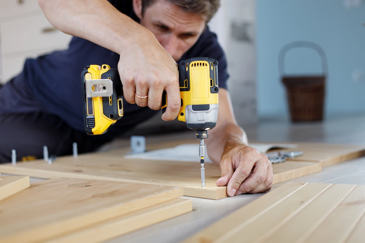 Intérieur d'atelier, homme en vêtement de travail bleu utilisant une perceuse-visseuse sans fil jaune et noire sur planche de bois clair