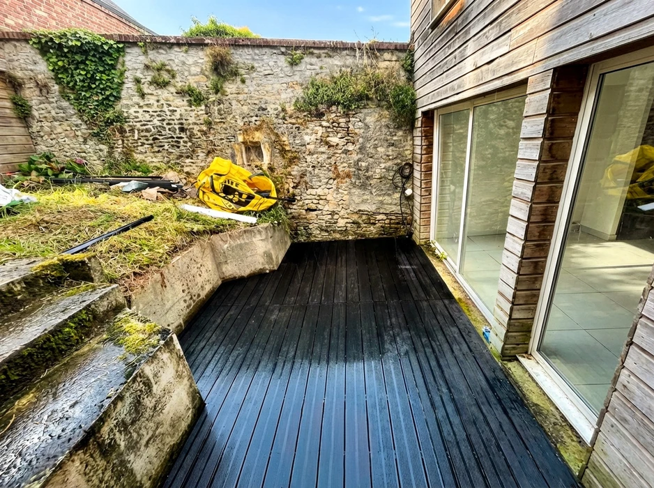 Terrasse extérieure en lame de bois composite teintée noir, adossée à un mur en pierre apparente ancien envahi de végétation